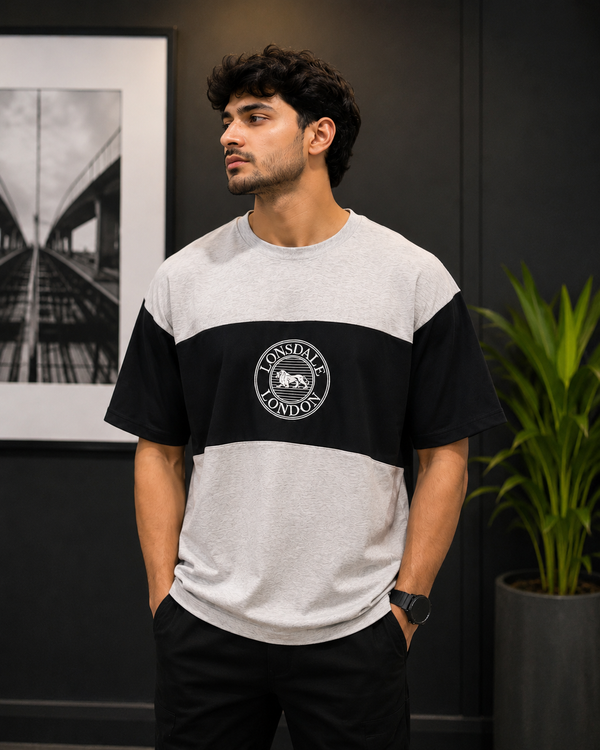 A man wearing a heather gray and black colorblock t-shirt with a circular 'Los Altos London' logo on the chest, standing in a modern room with a dark wall, framed picture, and a potted plant.