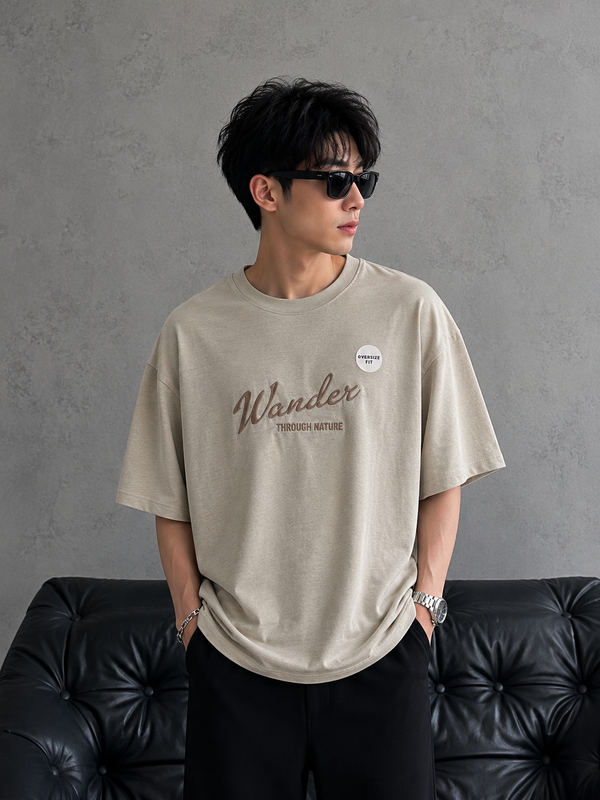 A man wearing an oversized beige graphic t-shirt with 'Wander' printed on the chest, paired with black pants and sunglasses, standing in front of a gray textured wall and a black leather tufted sofa.