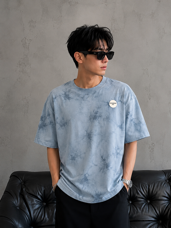 A man wearing a light blue tie-dye t-shirt with a small circular white logo on the chest, paired with black pants and sunglasses, standing in front of a textured gray wall and a black leather sofa.