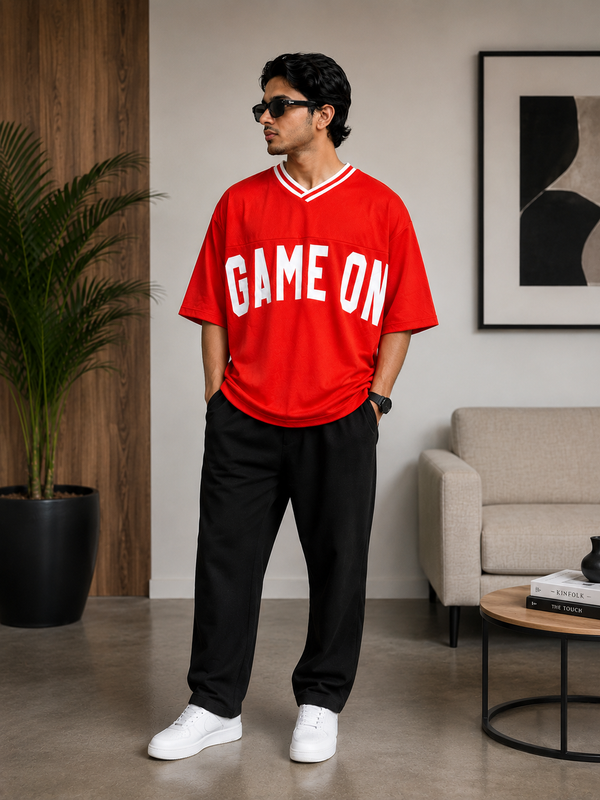 A man wearing a bright red v-neck t-shirt with 'GAME ON' printed in white across the chest, paired with black sweatpants and white sneakers, standing in a modern living room setting.