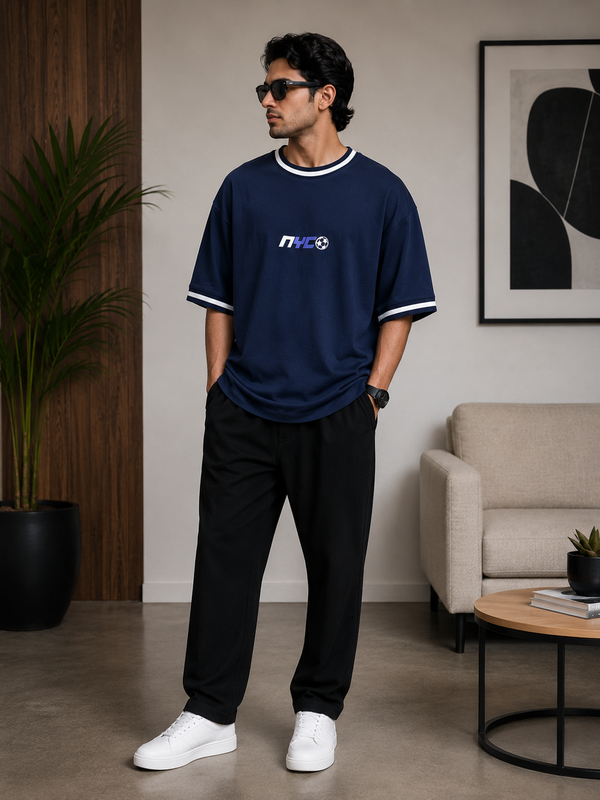 A man wearing a navy blue t-shirt with white trim and a white, blue, and black 'NFE3' graphic on the chest, paired with black pants and white sneakers, standing in a modern living room setting.