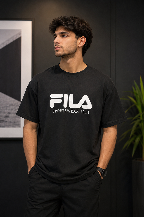 A man wearing a black FILA crewneck t-shirt with a bold white logo on the chest, standing in a dark indoor setting.