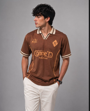A man wearing a brown short-sleeve polo shirt with cream and gold accents, paired with off-white trousers, standing against a solid gray studio background.