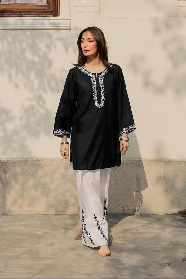 Woman wearing a black embroidered top and white pants standing against a light-colored wall.