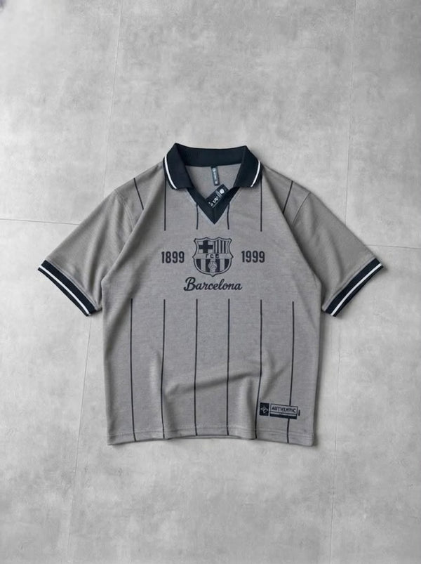 A light gray short-sleeve polo shirt with dark navy pinstripes, a dark navy collar, and 'FC Barcelona 1899 1999' printed on the chest, laid flat on a textured gray background.