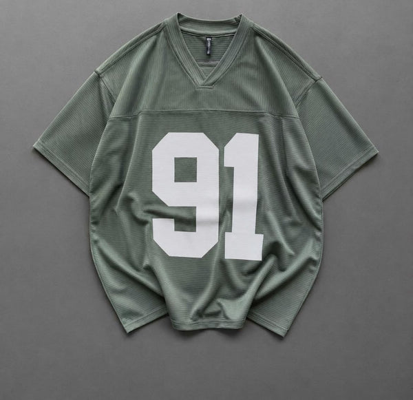 An oversized short-sleeve athletic jersey in sage green with a large white '91' printed on the front, laid flat on a dark gray background.